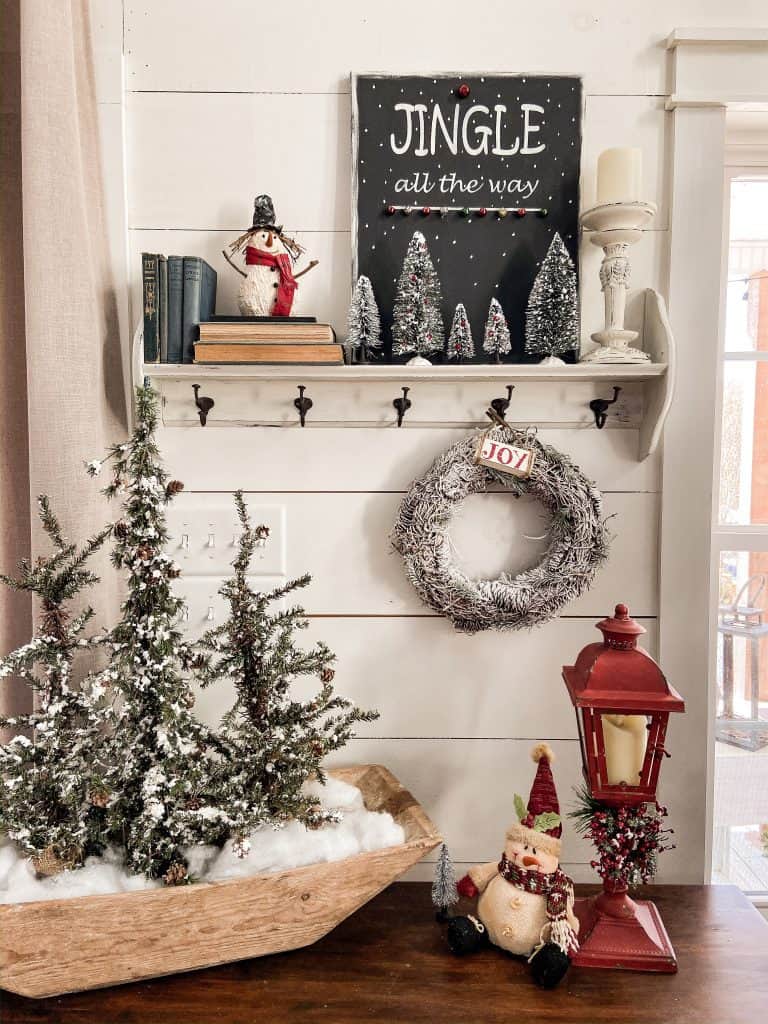 Entry Table Decorated for Christmas with vintage dough bowl filled with frosted trees, red lantern, snowman and shelf filled with bottle brush trees