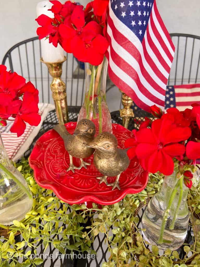 4th of July Centerpiece with brass birds and red geraniums and flags on a red cake plate.  