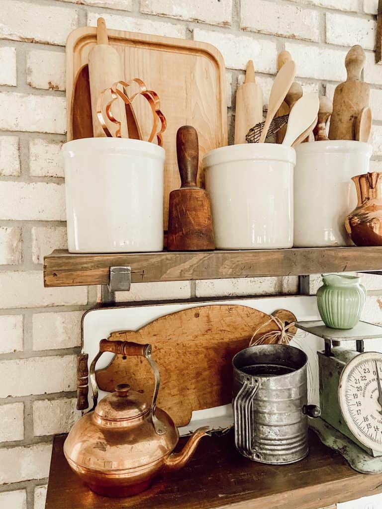 A collection of vintage crocks filled with vintage kitchen utensils on open shelves with a brick wall 