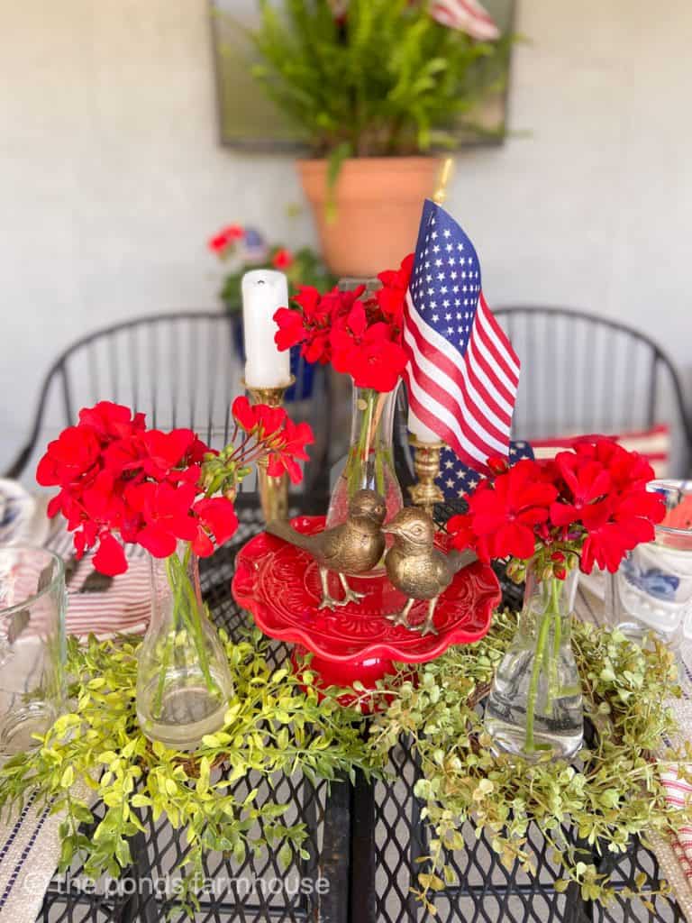 4th of July Centerpiece with brass birds and red geraniums and flags on a red cake plate.  