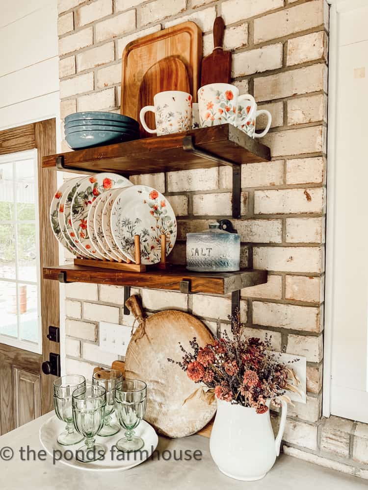 Open Shelf with dinnerware in festive colors. floral dinnerware and wooden bread boards.  Brick Wall.