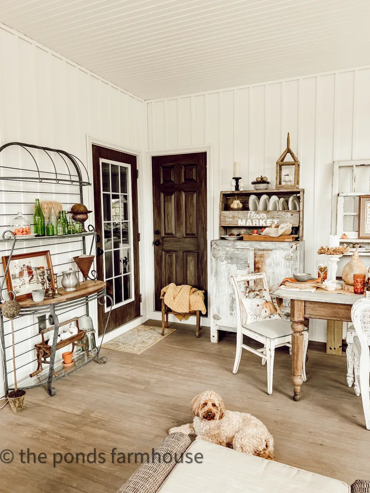 Dining area on screened porch with bakers rack and farm table.  Metal vintage icebox and fall decorations.  