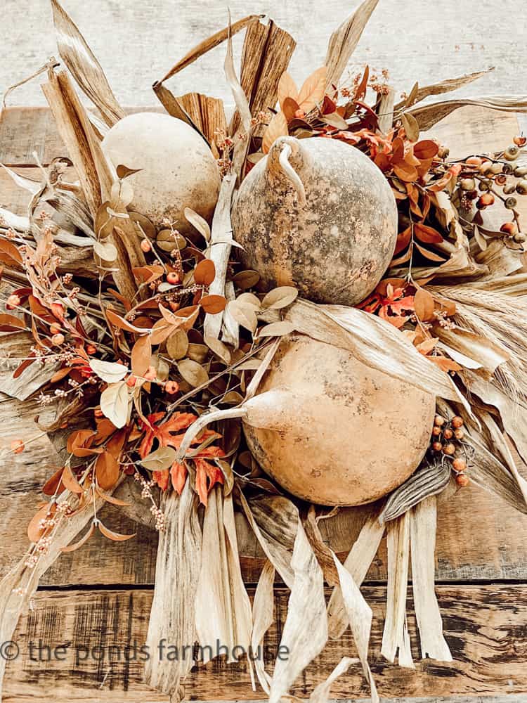 Gourdes, corn stalks, dried okra pods, berries and faux fall stems fill a wooden bowl on coffee table.