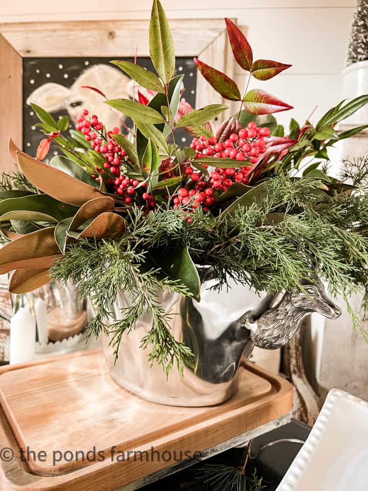 Pewter container filled with fresh greenery, cedar, magnolia leaves, and Nandina with red berries.