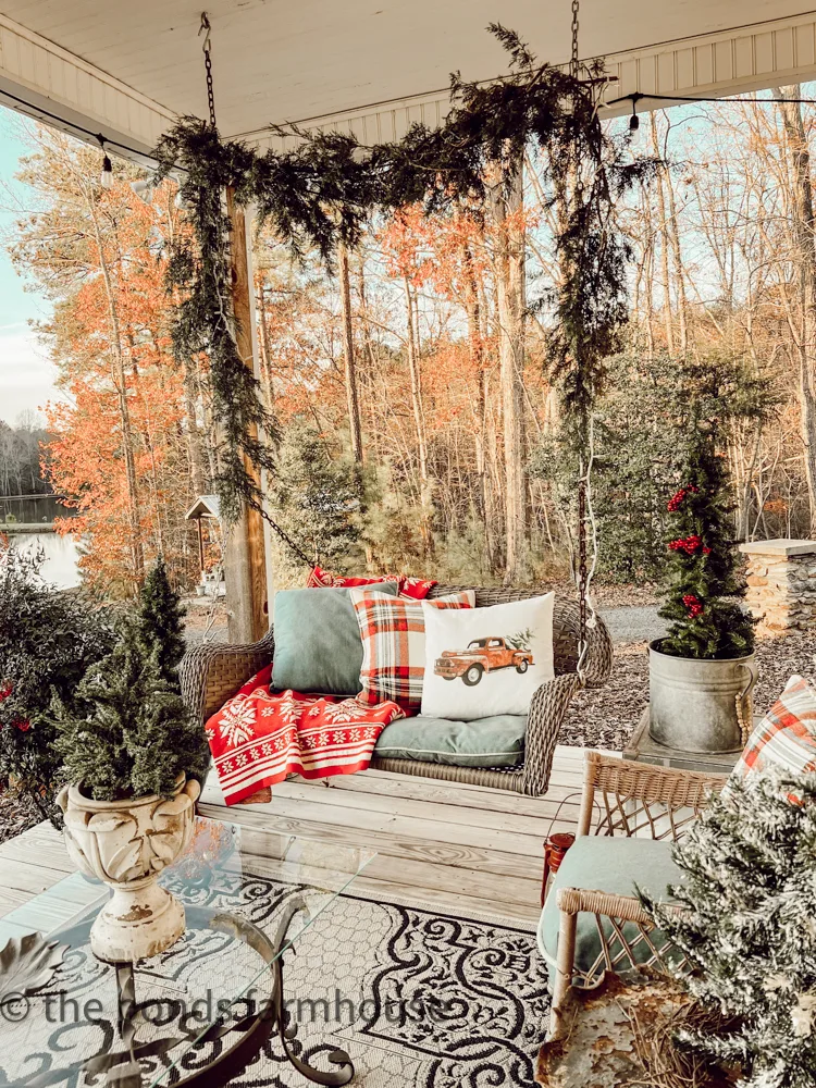 Farmhouse Front Porch Swing with holiday pillows and mini Christmas Trees.