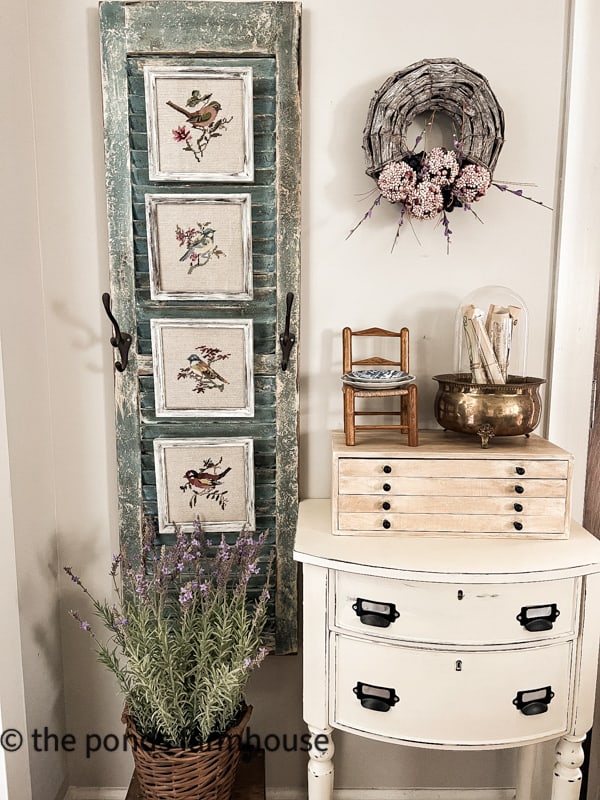 Hang thrifted needlepoint prints on a painted wooden shutter for a farmhouse style display.