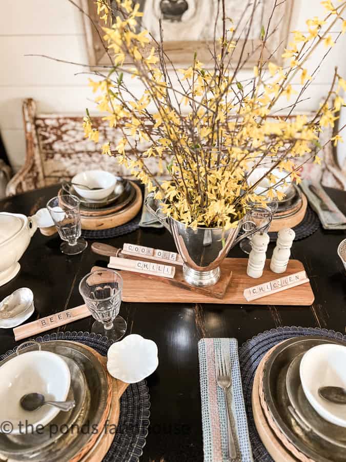 Silver urn holds forced yellow bell blooms with scrabble tiles and vintage bread boards for centerpiece.