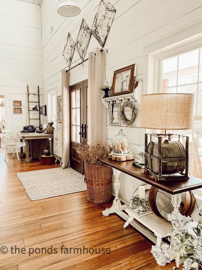 Repurposed gas can lamp on entry table with basket full of dried stems and pods beside front doors of farmhouse.