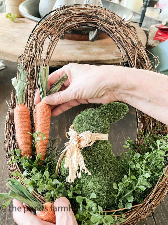 Add faux carrots to the Easter Wreath DIY.  Oval Wreath with artificial greenery, moss bunny and carrots.