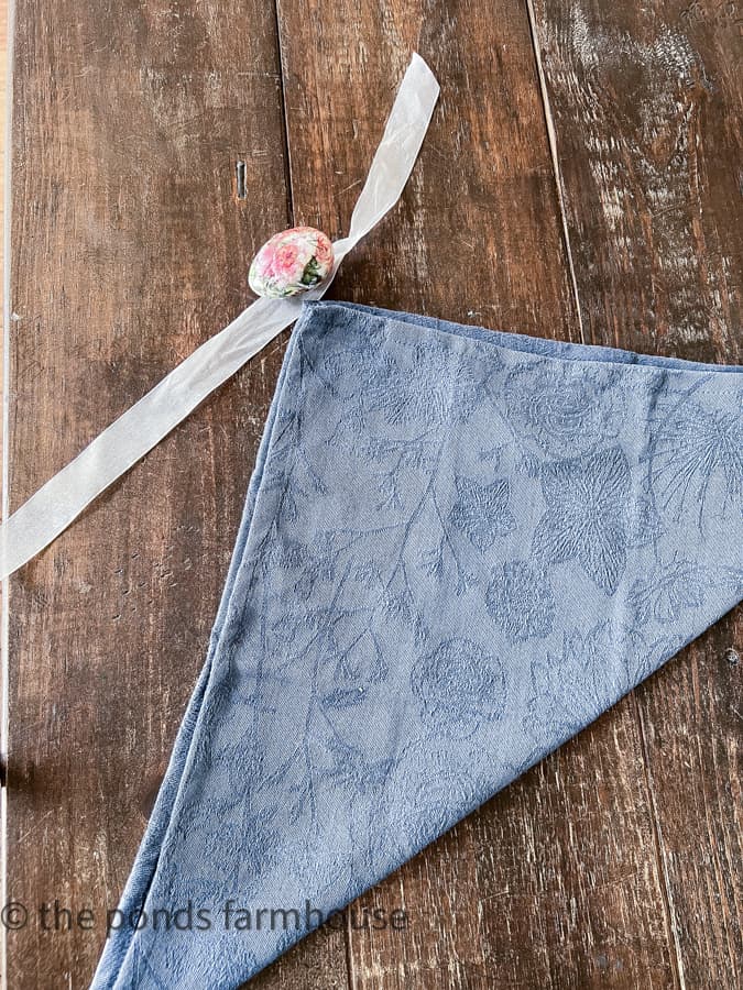 To Fold A Napkin Like Bunny Ears, fold a square napkin in half.  Blue cloth napkin with decoupage egg and white ribbon.