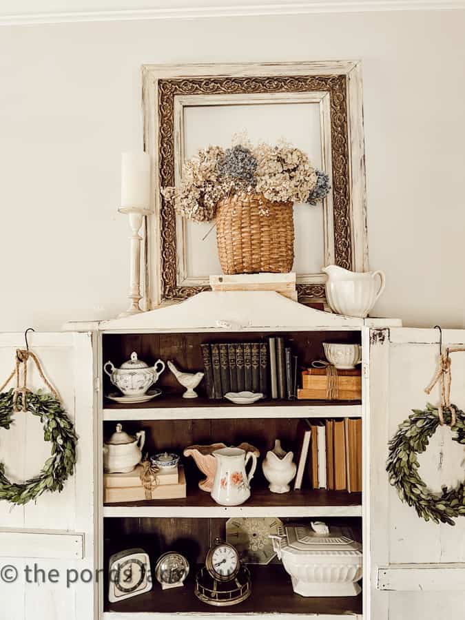 Dried flowers in old handmade basket framed by a vintage picture frame on top of a step back hutch in bedroom.