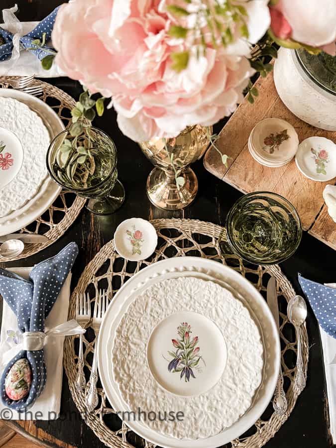 Table Easter Decorations include thrift store floral plates, green water goblets, woven placemats, and bunny ear napkins.