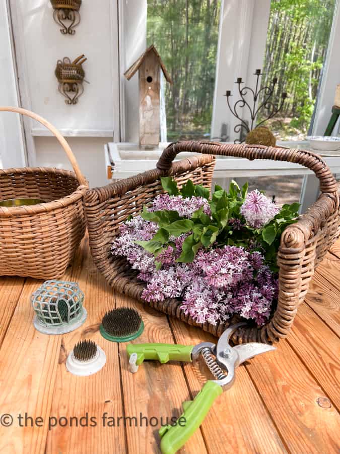 Harvest basket filled with fresh lilac blooms, old flower frogs and pruning shears. Basket Decor Ideas using gathering baskets.