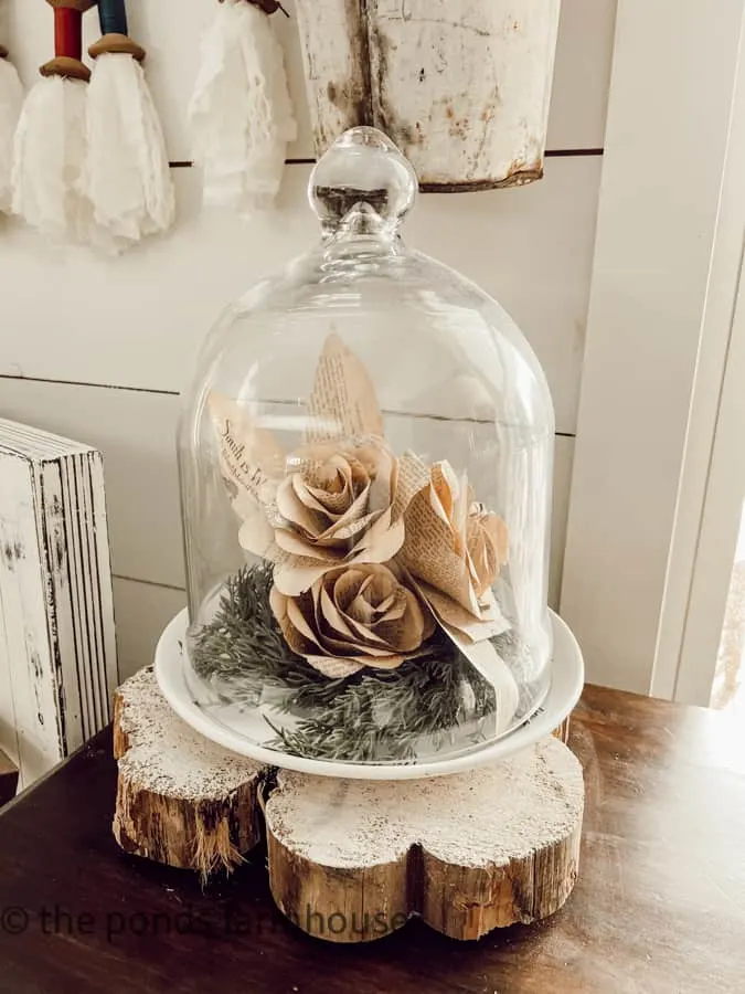 Paper roses under a glass dome sitting on a shallow white bowl.