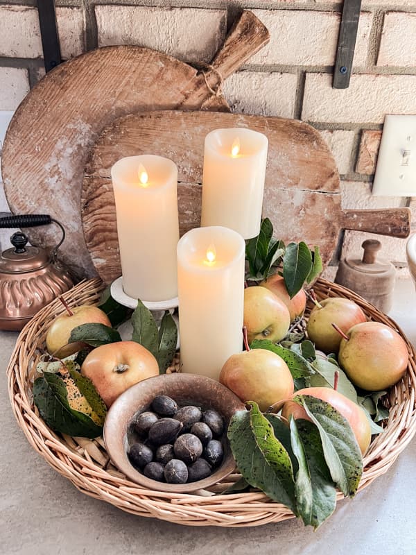 Woven tray with apples and foraged nuts with flicker candles.  Vintage breadboards stacked behind on brick wall.