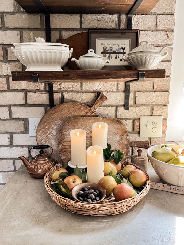 woven basket with apples and candles in front of bread boards below the open shelving.