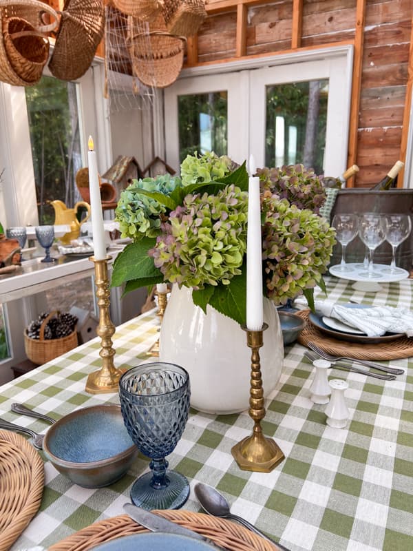 green and white buffalo check tablecloth with blue dishes and blue goblets, brass candlesticks and hydrangea arrangement.