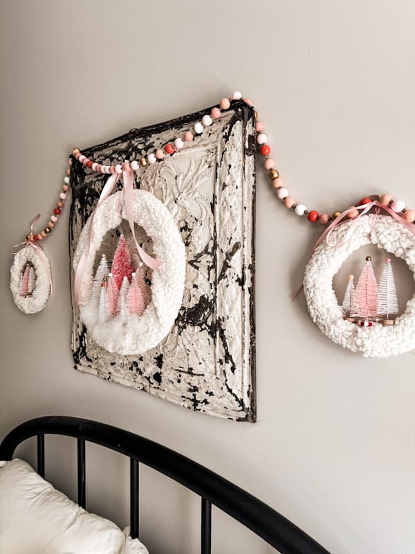 A trio of Easy Pink Bottle Brush Tree Wreaths hang on the bed on a vintage ceiling tin for a whimsical holiday look. 