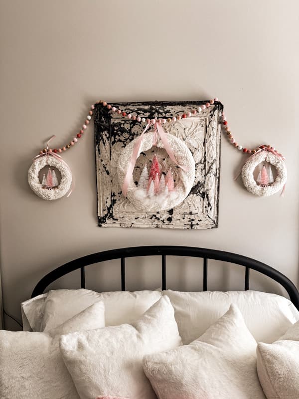 A trio of Pink Bottle Brush Tree Weaths on a antique ceiling tin with a black iron bed and snow white pillows.