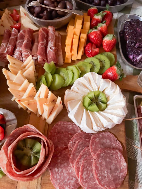 cheese, meats, goat cheese, olives, grapes, strawberries, kiwii, brie on a large wooden board for a grazing table.