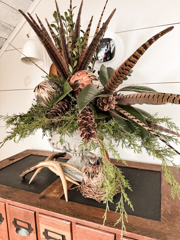 Pheasant feathers and ornament combine with pinecones and greenery for a table centerpiece.
