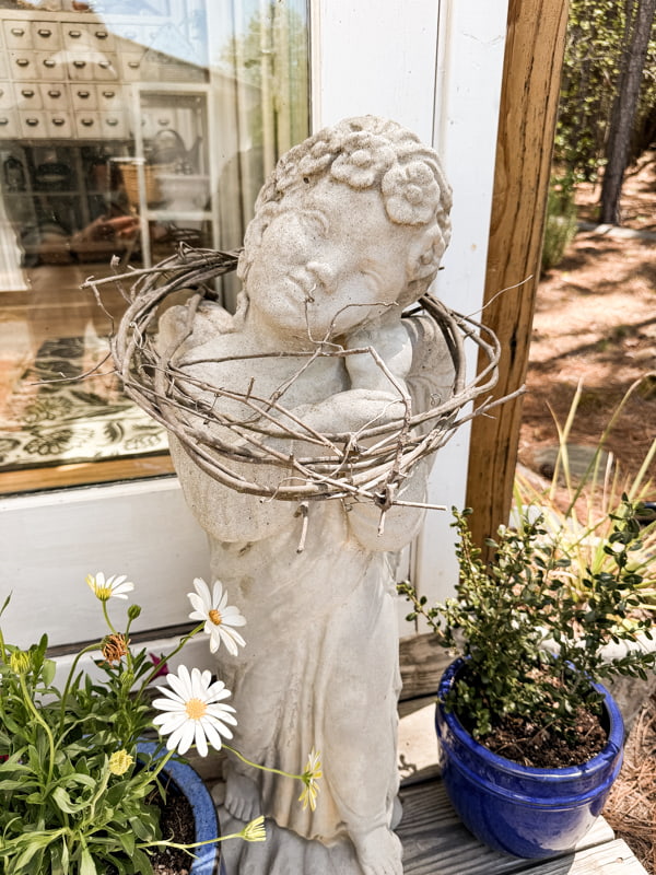 Vintage concrete garden angel with grapevine wreath and plants on Greenhouse front porch.