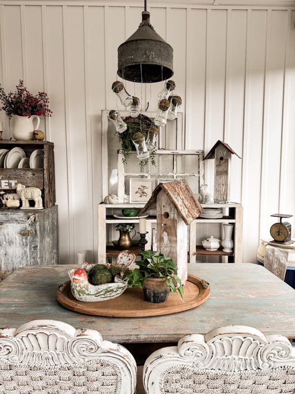 Centerpiece on farmhouse table with birdbath and DIY birdhouse with pot of ivy.  Moss balls in light fixture vases.  