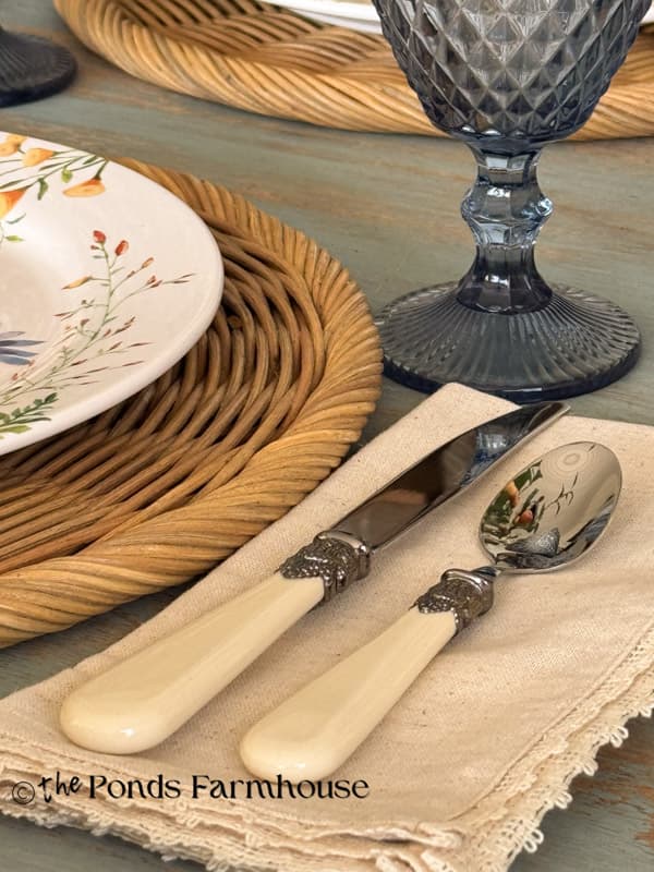 Bone silverware with elegant details on beige napkins beside the rattan placemats and blue goblets