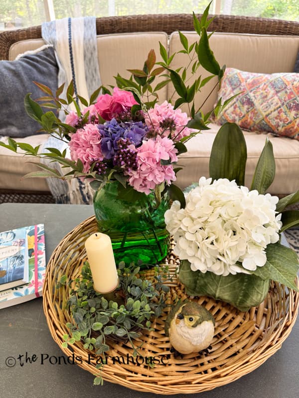 hydrangea arrangement in green glass & lettuce planter with brass candlestick and bird in round basket tray on coffee table on porch